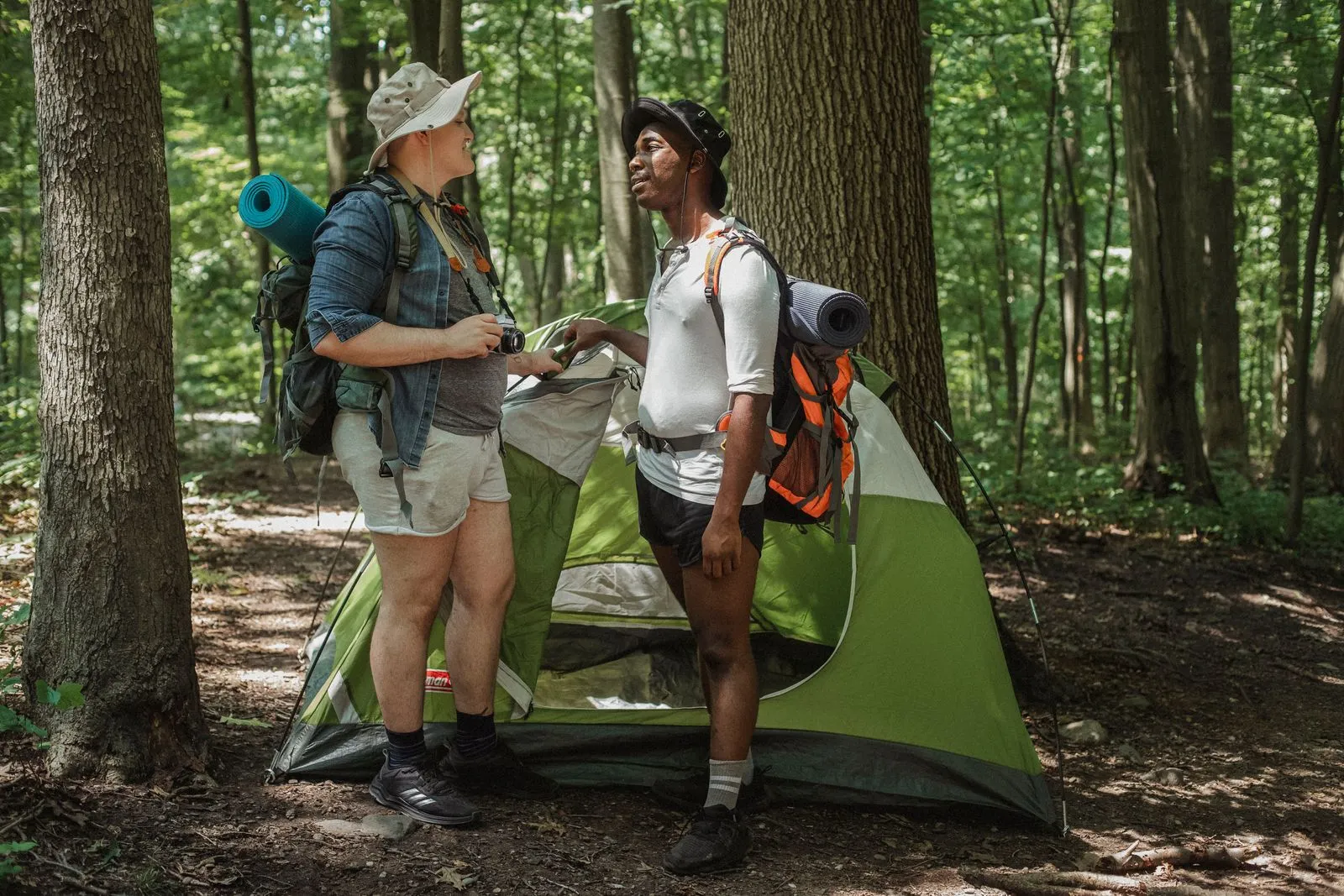 Full body of smiling multiethnic men traveling together in nature and chatting in leisure standing near tent in summer woods